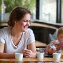 Two mothers smiling and drinking coffee with their toddlers nearby.